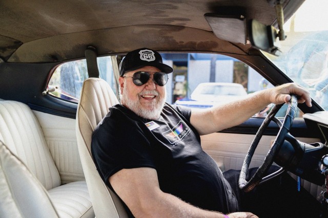 View from the passenger seat of a man sitting in the driver&#039;s seat, smiling at the camera, wearing a black hat, sunglasses and black shirt, with one hand on the steering wheel.