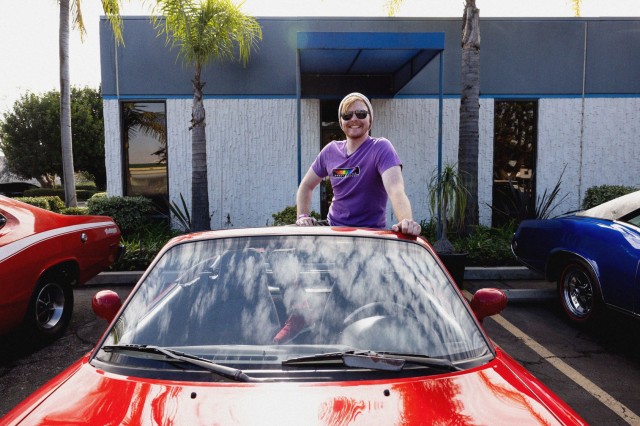 Man in sunglasses and purple t-shirt standing up in the driver seat of his red convertible Miata.