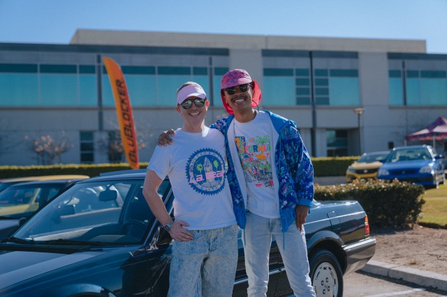 Two men dressed in 1980s fashion pose together, arms across each others&#039; shoulder, in front of a car show