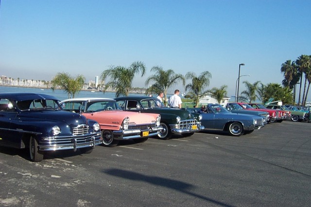 Classic cars lined up in a parking lot next to the ocean with the city of Long Beach in the background.