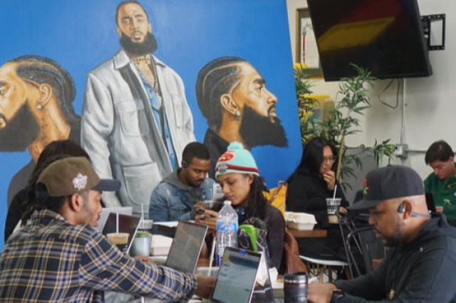 A group of people sitting at black circular tables inside South LA Cafe, working on laptops. In the background, a large, vibrant blue mural of Nipsey Hussle watches over the room. The space is filled with a sense of quiet focus and community connection.