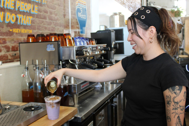 Asian woman in black t-shirt and black bandana preparing an iced coffee drink at a coffee shop.