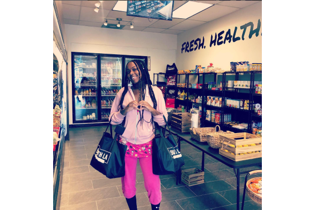 A young woman standing in a small grocery market, carrying two grocery bags.