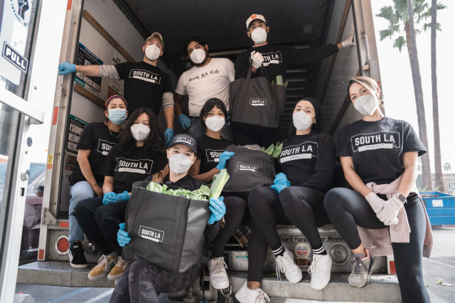 A group of ten South LA Cafe volunteers, wearing protective face masks, gloves, and branded black SLAC shirts, pose together in the back of a delivery truck. They are holding large black reusable grocery bags filled with fresh leafy greens and produce, ready for distribution to the community.