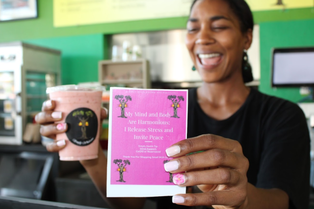 A joyful Mia Keeling laughs while holding a pink smoothie in one hand and a pink affirmation card in the other. The card reads: "My Mind and Body Are Harmonious; I Release Stress and Invite Peace."