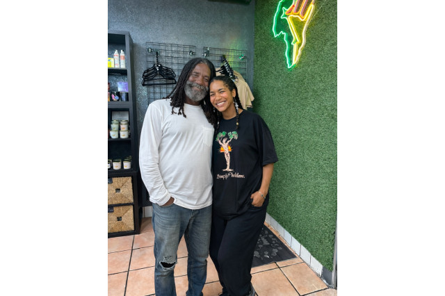 A smiling man and woman stand close together indoors. The man has long dreadlocks, a grey-flecked beard, and wears a white long-sleeve shirt with distressed jeans. The woman has braided hair and wears a black graphic T-shirt. They are positioned in front of a green textured wall featuring a neon light.