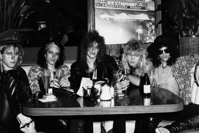 Black and white photograph of the young 1980s rock band, Guns n' Roses, sitting at a restaurant booth together.