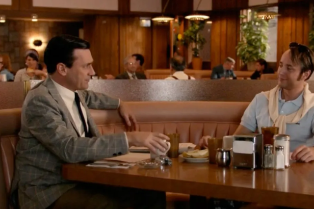 Man holding a photo from a TV show of two men in a booth, one in a suit and one in a tennis outfit.