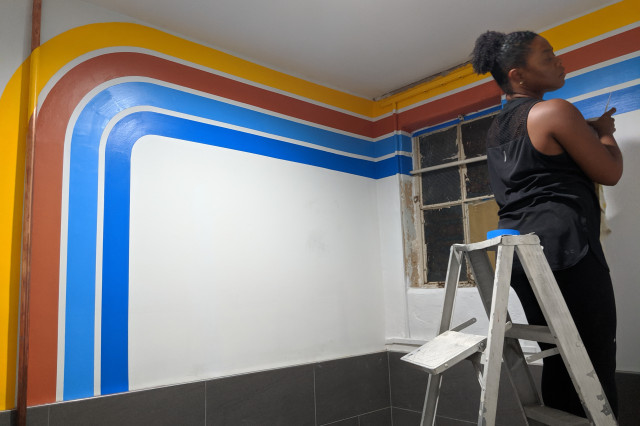 A woman sits atop a ladder painting blue lines on a wall with yellow, orange and lighter blue lines along a top of a wall. 