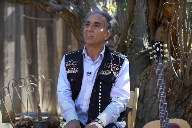 Man with white shirt and black vest. Guitar at side