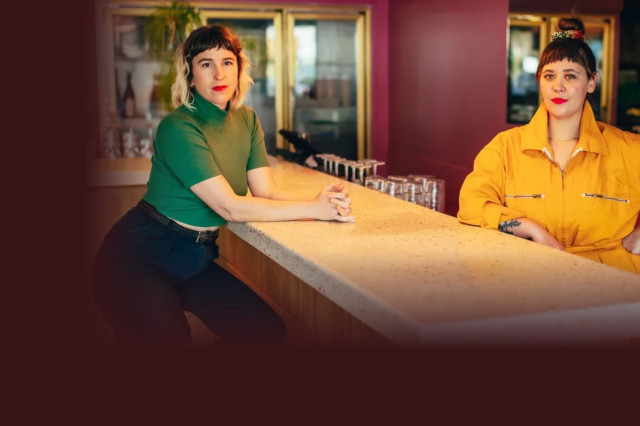 two people pose behind a long counter inside The Ruby Fruit. One wears a green top and dark pants, while the other wears a bright yellow jumpsuit. In the foreground, a lime green graphic banner features the bar's name next to an image of a red cocktail garnished with a dehydrated citrus wheel.