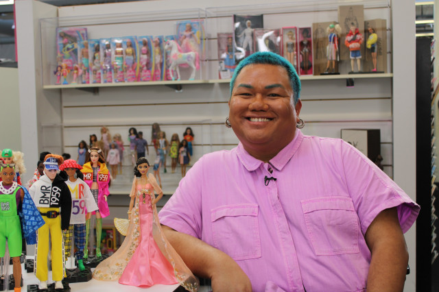 Man wearing pink jumper with several barbie dolls next to him and in the background