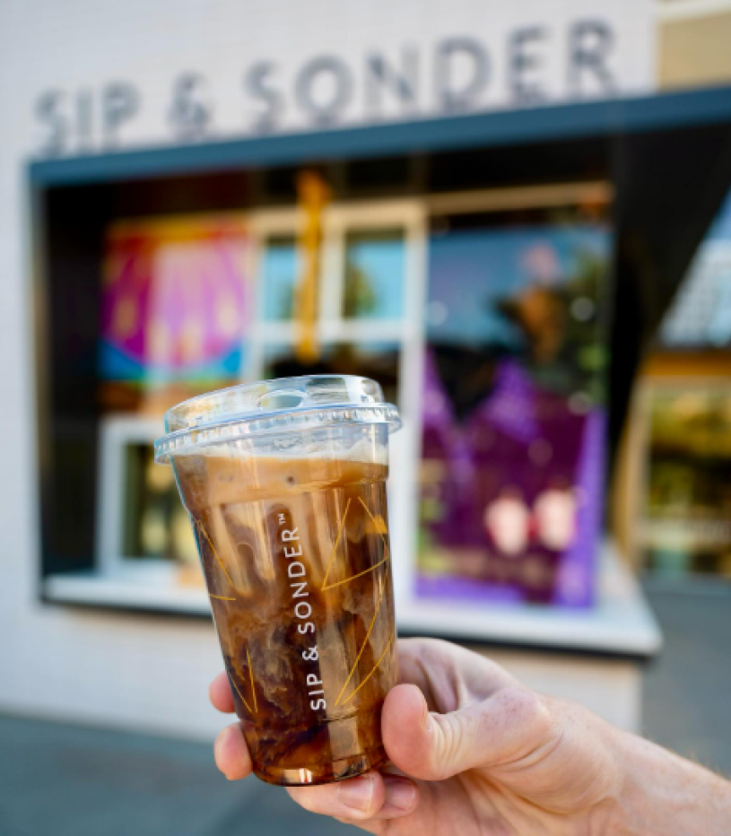 A hand holds up a cup of coffee with a logo that reads "Sip & Sonder" in front of a coffee shop labeled the same.