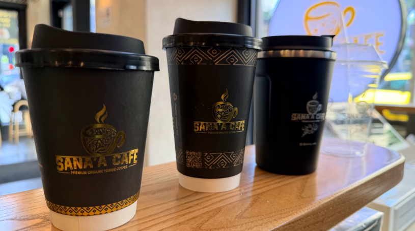 Three black takeaway coffee cups with lids, branded “Sana'a Cafe,” sit on a wooden counter inside a café, with a window and blurred street scene in the background.